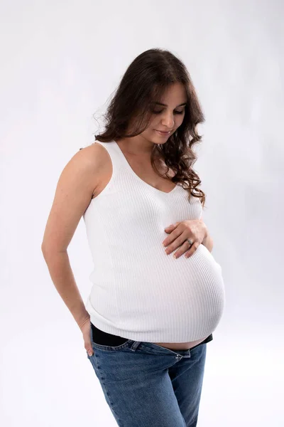 Pregnant brunette woman with long hair. Expectant mother gently touches her tummy, looks down. Studio light, white background. Concept of waiting for a baby, healthy pregnancy, childbearing age