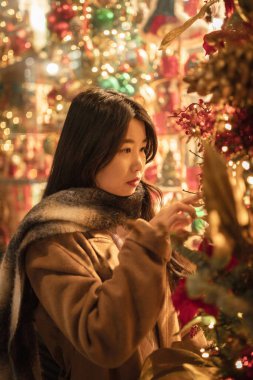 Eşarba sarılmış Asyalı bir kadın, profilde duruyor, güzel vitrinlere bakıyor, Noel, kış. Çinli, mutlu esmer kız. Tatil, alışveriş, hediyeler, dekor konsepti. Avrupa şehri.