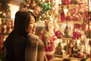 Eşarba sarılmış Asyalı bir kadın, profilde duruyor, güzel vitrinlere bakıyor, Noel, kış. Çinli, mutlu esmer kız. Tatil, alışveriş, hediyeler, dekor konsepti. Avrupa şehri.