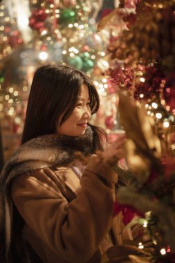 Eşarba sarılmış Asyalı bir kadın, profilde duruyor, güzel vitrinlere bakıyor, Noel, kış. Çinli, mutlu esmer kız. Tatil, alışveriş, hediyeler, dekor konsepti. Avrupa şehri.