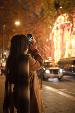 Cep telefonu kullanan Asyalı bir kız, sokakta dikilip gece lambalarının fotoğrafını çekiyor. Uzun saçlı Çinli kadın, Avrupa 'yı geziyor, şehirde yürüyor. Kavram turizmi, blog yazma, internet