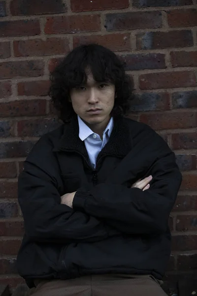 Japanese guy, stylish outfit, sitting with crossed arms against background of brown bricks wall, daylight, dark tones. Asian man 25 years old with long curly hair. Concept of fashion, self-confident, loneliness.
