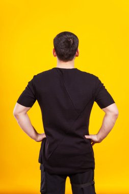 Sarı arka planda siyah tişörtlü bir adam. Siyah tişörtlü, sade bir arka planı olan bir adamın arkası. Bir fotoğraf stüdyosunda yaz kıyafetlerinin fotoğraf çekimi.
