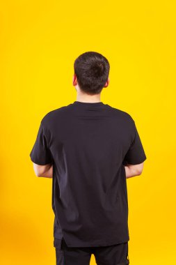 Sarı arka planda siyah tişörtlü bir adam. Siyah tişörtlü, sade bir arka planı olan bir adamın arkası. Bir fotoğraf stüdyosunda yaz kıyafetlerinin fotoğraf çekimi.