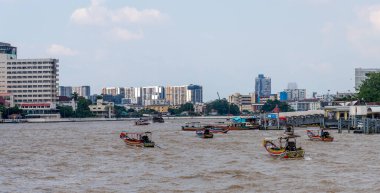 Bulanık sularda yüzen renkli teknelerin olduğu hareketli bir nehir manzarası, arka planda bir şehir silüeti var..