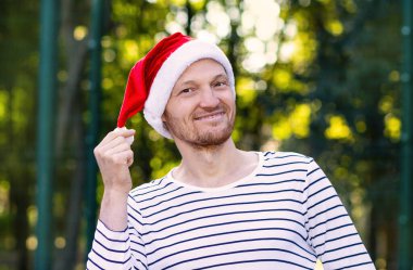Genç yakışıklı adam Noel Baba şapkası takıyor. Noel Baba şapkalı gülümseyen sakallı bir adamın portresi. Noel 'de yakışıklı bir genç. Noel Baba 'nın yakın çekimi. Çizgili tişörtlü bir adam.