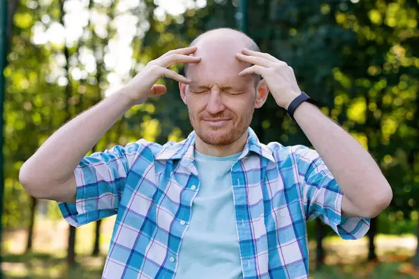 The man has a headache. A bald man is resting in nature. A man thinks in close-up. Portrait of a guy with his hands on his head. Stress in a business person. Migraine from overwork