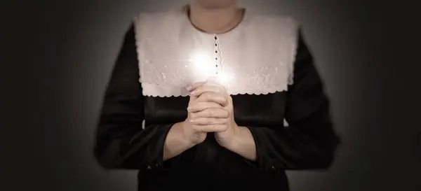 stock image A nun dressed in traditional attire is seen praying with her hands clasped together, emanating a soft glowing light. This image represents faith, spirituality, and devotion.