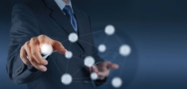 Бизнесмен в костюме взаимодействует с футуристическим виртуальным интерфейсом. Его палец направлен на светящийся узел, представляющий цифровую связь и бизнес-технологии..