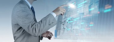 Gri takım elbiseli bir işadamı, karmaşık veriler, çizelgeler ve çizelgeler gösteren fütüristik bir dijital arayüzle etkileşime geçiyor. Bu resim, işletme yönetimi için yapay zekanın kullanımını vurgular.