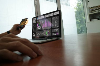 Akıllı bir telefondaki finansal grafikleri ve modern bir ofisteki veri paneli olan bir dizüstü bilgisayarı analiz eden kişi.