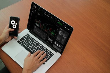  Finansal ticaret verileri ve otomasyon simgelerini gösteren dijital arayüzlü bir dizüstü bilgisayar ve akıllı telefon kullanan ellerin yakın görüntüsü.