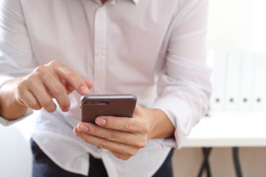  Close up of man in white shirt holding and using smartphone with both hands indoors. Concept of communication, technology, social media, mobile app, texting, and online business activities.