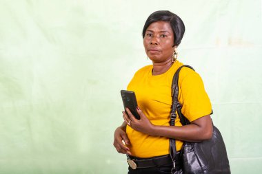 Sarı tişörtlü olgun bir kadın yeşil arka planda cep telefonuyla durup düşünürken.