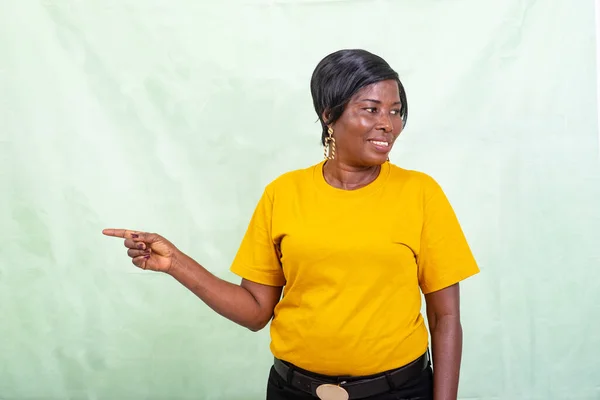 stock image beautiful mature woman in yellow t-shirt standing over green background pointing finger sideways and looking the other side smiling.