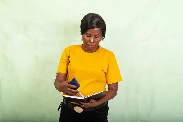 携帯電話を持ちながら日記をつけた緑の背景に立つ黄色のTシャツを着た美しい熟女 — ストック写真