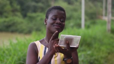 Genç, şık, Afrikalı bir kadın parkta durmuş küçük bir aynada yüzüne bakıp gülümsüyor..