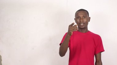 Kırmızı tişörtlü yakışıklı bir adam beyaz arka planda dikilip kameraya gülümserken jestlerle konuşuyor..