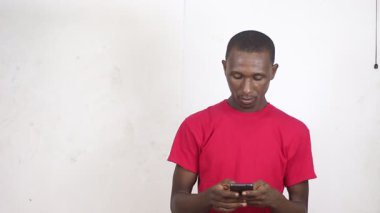 Kırmızı tişörtlü yakışıklı bir adam beyaz bir arka planda cep telefonuyla zafer jesti yaparak gülüyor..