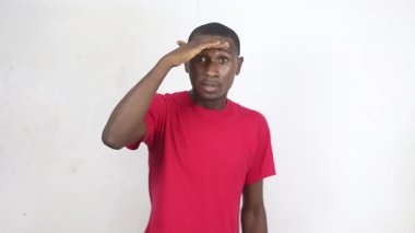 Kırmızı tişörtlü yakışıklı adam beyaz arka planda duruyor elini alnına koyuyor iyi görmeye çalışıyor ve güneş gözlüğü takıyor gülümsüyor.