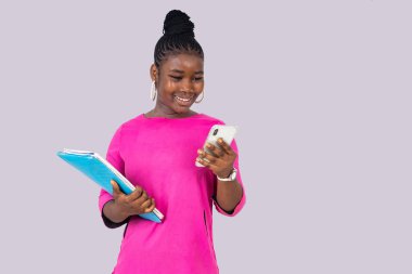 Beyaz arka planda duran pembe gecelikli güzel bir kız öğrenci elinde cep telefonu ve defterleriyle gülümsüyor..