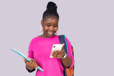 Beyaz arka planda duran pembe gecelikli güzel bir kız öğrenci elinde cep telefonu ve defterleriyle gülümsüyor..