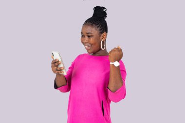 Pembe paçalı güzel bir kız öğrenci elinde cep telefonuyla beyaz arka planda durup gülerken zafer işareti yapıyor..