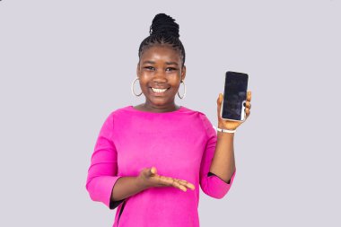 Pembe paçalı güzel bir kız öğrenci beyaz arka planda durup gülerken cep telefonunu gösteriyor..