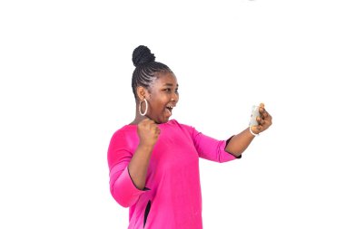 Pembe paçalı genç bayan öğrenci beyaz arka planda duruyor cep telefonu ekranına bakıyor ve zafer işareti olarak yumruğunu kaldırıyor..