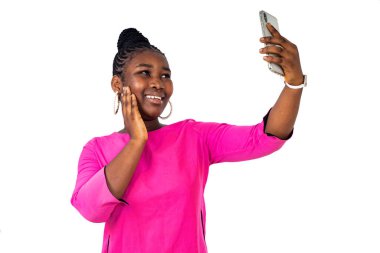 Beyaz arka planda duran pembe gecelikli genç bir kız gülümserken cep telefonuyla video görüşmesi yapıyor..