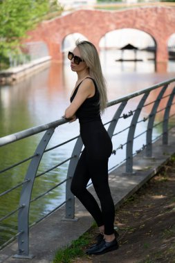 Güneş gözlüklü genç sarışın bir kadın şehir parkındaki bir göletin kıyısında duruyor. Kız bir şey düşündü, doğayı düşündü, zihinsel olarak dinleniyor. Yüksek kalite fotoğraf