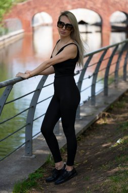 Güneş gözlüklü genç sarışın bir kadın şehir parkındaki bir göletin kıyısında duruyor. Kız kameraya bakıyor. Yüksek kalite fotoğraf