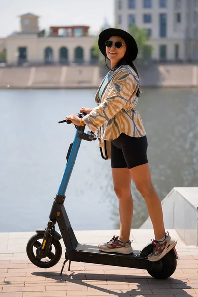 Güneş gözlüklü ve şapkalı genç bir Asyalı kadın scooterın üzerinde duruyor. Seddin üzerindeki bir kızın yaz şehir portresi. Yüksek kalite fotoğraf
