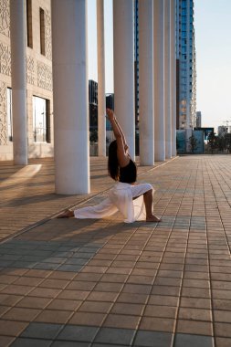 Genç Asyalı kadın gündüz vakti şehir manzarasıyla yoga yapıyor. Kız binanın sütunlarına bakarak asanas gösterisi sergiliyor. Şehir yogası, gün doğumu egzersizi..