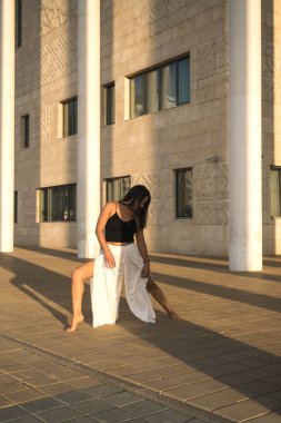 Genç Asyalı kadın gündüz vakti şehir manzarasıyla yoga yapıyor. Kız binanın sütunlarına bakarak asanas gösterisi sergiliyor. Şehir yogası, gün doğumu egzersizi..