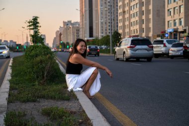 Yüzünde gülümseme olan Asyalı bir kız yolun kenarında oturuyor. Yazın otostop yapmak..