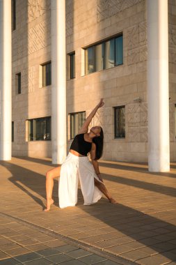 Genç Asyalı kadın gündüz vakti şehir manzarasıyla yoga yapıyor. Kız binanın sütunlarına bakarak asanas gösterisi sergiliyor. Şehir yogası, gün doğumu egzersizi..