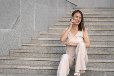 Genç bir kadın dışarıdaki merdivenlerde oturuyor ve cep telefonuyla konuşuyor. Yüksek kalite fotoğraf