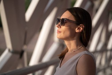 Bej elbiseli ve güneş gözlüklü esmer bir kadının yaz portresi. Şehir soyut arka planı, doğal ışığın güzel kırılması. Yüksek kalite fotoğraf