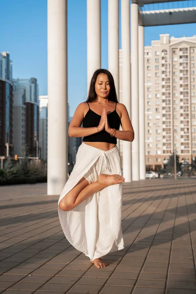Young Asian woman practicing yoga outdoor during daytime with the view of the city. The girl performs asanas with the view of the blue sky. Urban yoga, sunrise workout. Urban yoga, sunrise workout.