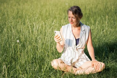 Genç sarışın bir kadın Lotus pozisyonunda bir vadide oturuyor ve akıllı telefondan mesajlar gönderiyor. Yüksek kalite fotoğraf