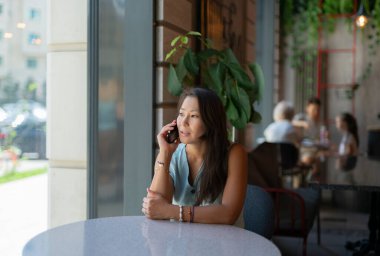 Genç bir Asyalı kadın bir kafede bir masada oturuyor ve cep telefonuyla konuşuyor. Yüksek kalite fotoğraf