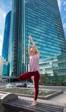 59 yaşında yetişkin bir kadının portresi. Şehir caddesinde yoga yapıyor. Kadının arkasında mavi bir gökdelen var. Yetişkinlerin aktif spor yaşam tarzı. 