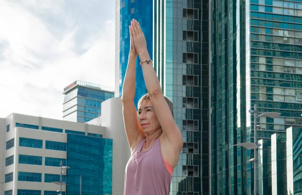 Portrait of a mature woman, 59 years old, doing yoga on a city street. Behind the woman is a blue skyscraper. Active sport lifestyle of mature people.