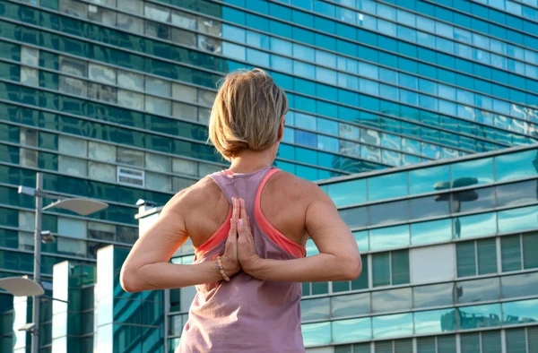 Portrait of a mature woman, 59 years old, doing yoga on a city street. In front of the woman is a blue skyscraper. Active sport lifestyle of mature people. High quality photo