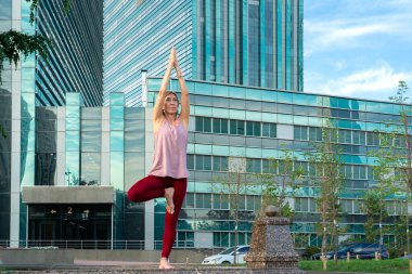 59 yaşında yetişkin bir kadının portresi. Şehir caddesinde yoga yapıyor. Kadının arkasında mavi bir gökdelen var. Yetişkinlerin aktif spor yaşam tarzı. 
