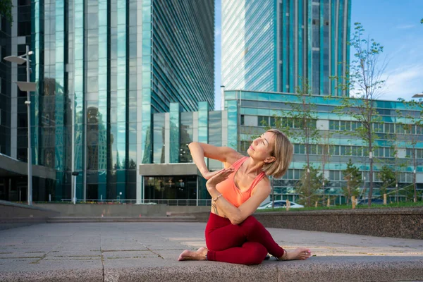 Portrait of a mature woman, 59 years old, doing yoga on a city street. Behind the woman is a blue skyscraper. Active sport lifestyle of mature people.