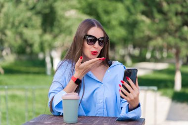 Uzun sarı saçlı bir kızın yaz portresi. Mavi tişörtlü ve güneş gözlüklü bir kız elinde bir akıllı telefon tutuyor ve video görüşmesiyle konuşuyor. Yüksek kalite fotoğraf