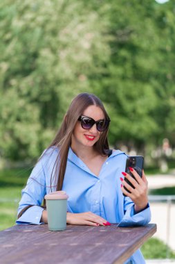 Uzun sarı saçlı bir kızın yaz portresi. Mavi tişörtlü ve güneş gözlüklü bir kız elinde bir akıllı telefon tutuyor ve video görüşmesiyle konuşuyor. Yüksek kalite fotoğraf