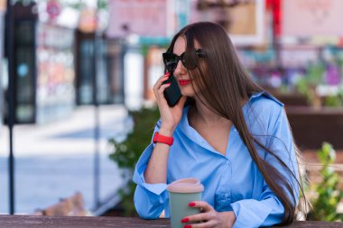 Uzun sarı saçlı bir kızın yaz portresi. Mavi gömlekli ve güneş gözlüklü bir kız akıllı telefondan konuşuyor. Yüksek kalite fotoğraf
